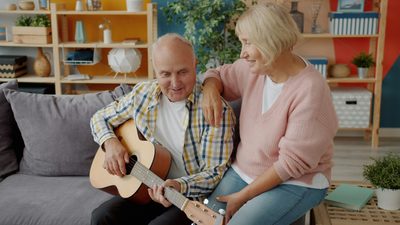 Mann lernt Gitarre, Frau sitzt unterstützend daneben